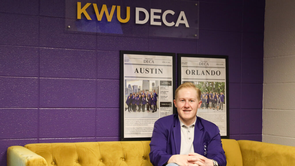 Man sitting on couch in front of KWU DECA sign and press clippings