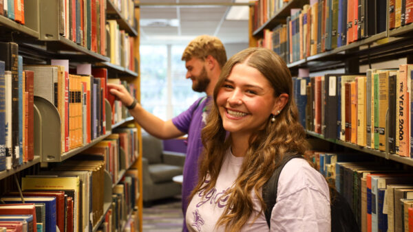 Student standing between library book stacks; another student looks through books behind her